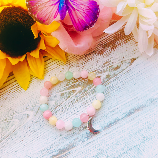 A silver-plated crescent moonstone quartz bracelet with polished beads, displayed on a wooden surface with colorful flowers and a butterfly in the background.